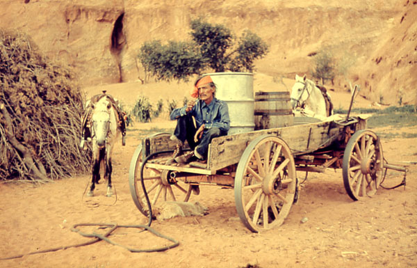 Navajo Man sitting in wagon, two barrels in the back of wagon, two horses on the side of the wagon